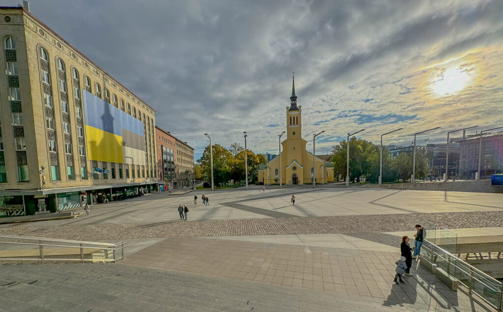 This photo shows Freedom Square (Vabaduse väljak) in Tallinn, Estonia, with the yellow St. John's Church (Jaani kirik) prominently visible on the right side. St. John's Church is a Lutheran church built in a neo-Gothic style in the 1860s, making it one of the oldest functioning churches in the city. The building on the left with the large flags is part of the modern development surrounding Freedom Square, which often displays art or political messages. Freedom Square itself is a significant public space in Tallinn, serving as a venue for events, celebrations, and gatherings, with historical monuments and symbolic importance tied to Estonia's independence.