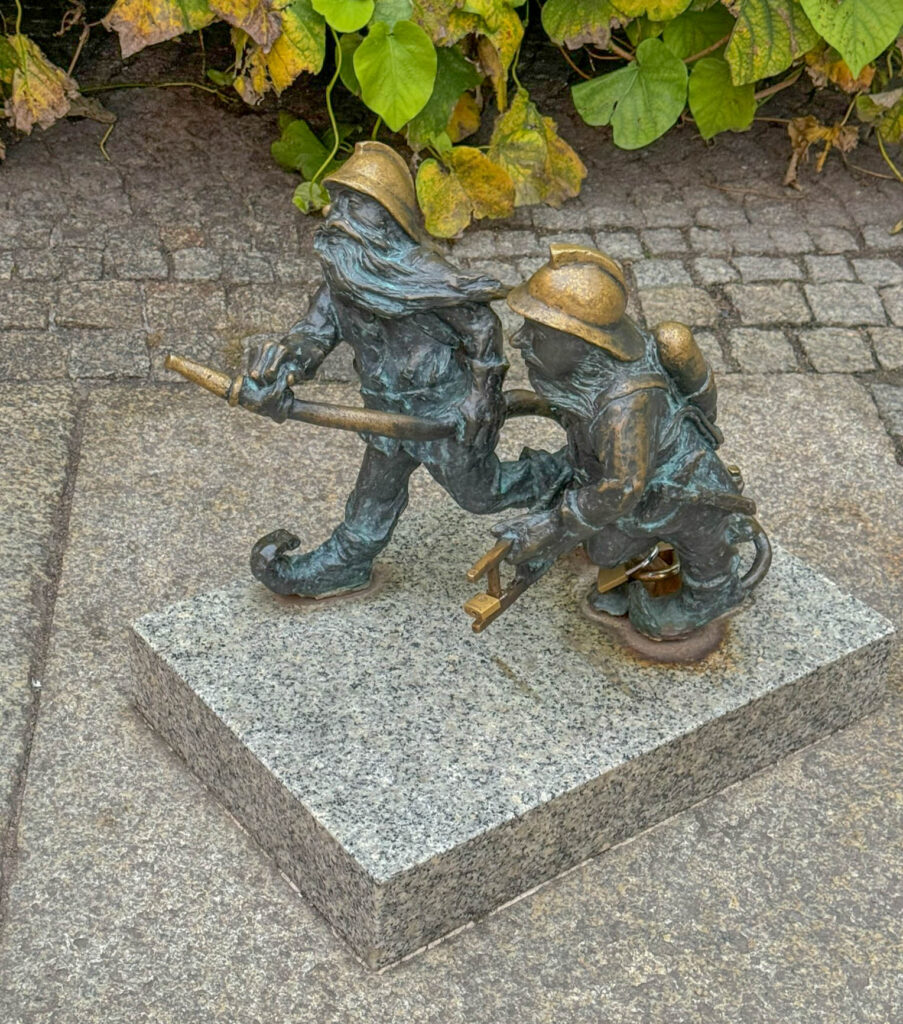 Here's a little sculpture called "Firefighter Gnomes" (Strażacy Krasnale), created as a heartfelt nod to firefighters. The scene features two gnomes decked out in firefighter helmets, gripping a hose, ready to take on imaginary flames. It’s a reminder of the courage and commitment that real-life firefighters bring to the job every day. These gnomes are part of Wrocław’s famous krasnale collection - tiny dwarf statues sprinkled all over the city. Locals and visitors alike enjoy hunting them down like a city-wide scavenger hunt. Each gnome tells its own story, often highlighting a piece of Wrocław’s history, culture, or the everyday heroes that keep the city running.