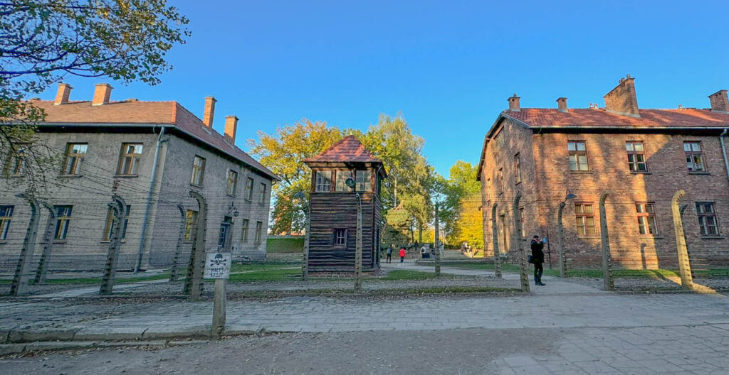 This image shows Auschwitz I, featuring a guard tower, prisoner barracks, and barbed-wire fencing. The guard tower was used for surveillance to prevent escapes, while the buildings housed prisoners and administrative offices. The warning sign with a skull and crossbones indicates the presence of an electrified fence, which was a deadly barrier around the camp.