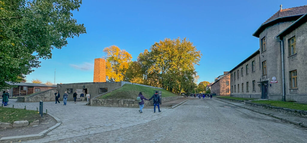 Here's a view of the gas chamber and crematorium at Auschwitz I from the opposite side, featuring the low, bunker-like structure with a brick chimney used for incineration. The sloped, grass-covered roof was designed to blend into the surroundings.