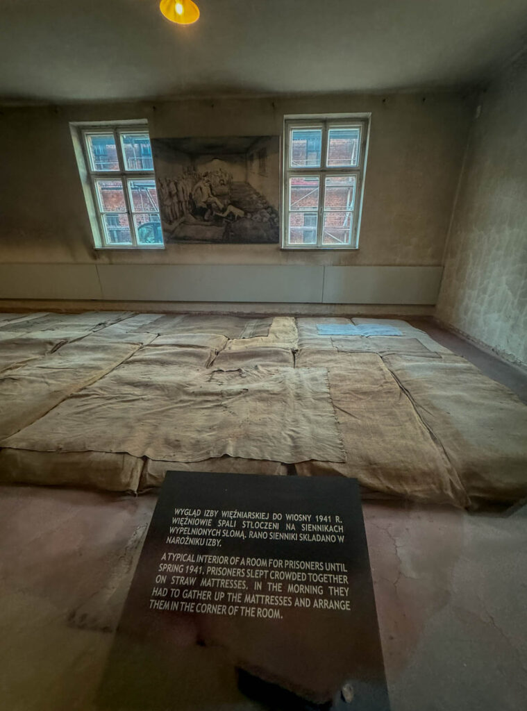 This image depicts the interior of a prisoner barracks at Auschwitz I, showing a reconstruction of the harsh living conditions. The floor is covered with straw-filled mattresses, where prisoners were forced to sleep crowded together. A sign explains that in the morning, inmates had to gather and stack the mattresses in a corner.