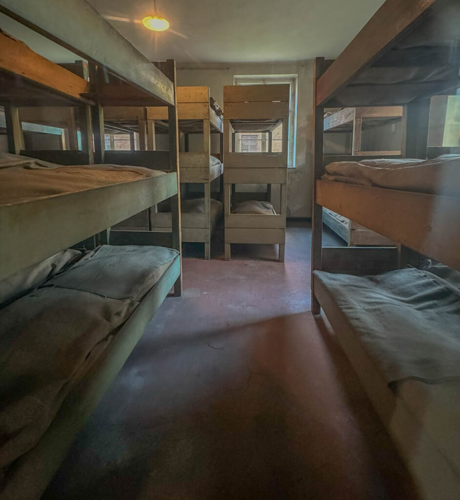 This image depicts the prisoner barracks at Auschwitz I, showing tightly packed wooden bunk beds where inmates were forced to sleep in harsh conditions. The triple-tiered bunks, with thin straw mattresses, were overcrowded, often holding multiple prisoners per level.