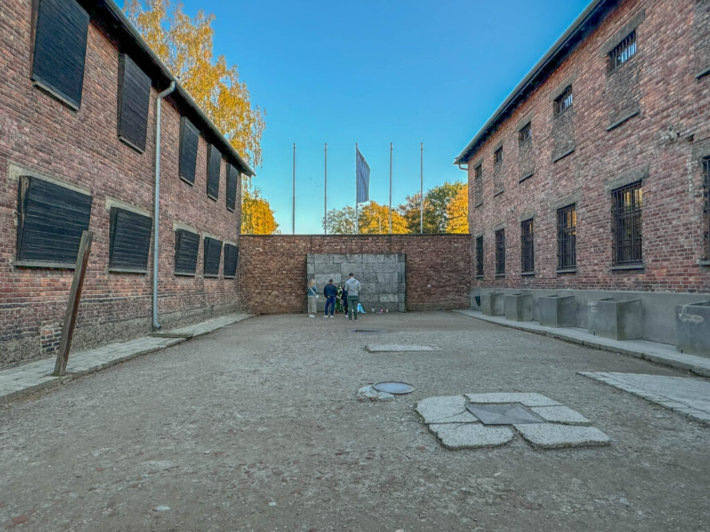 This image shows the execution courtyard at Auschwitz I, where prisoners were executed against the "Death Wall" between Blocks 10 and 11. The black-shuttered windows on the left were covered to prevent other inmates from witnessing the killings, while the concrete posts on the right were used for prisoner punishments.