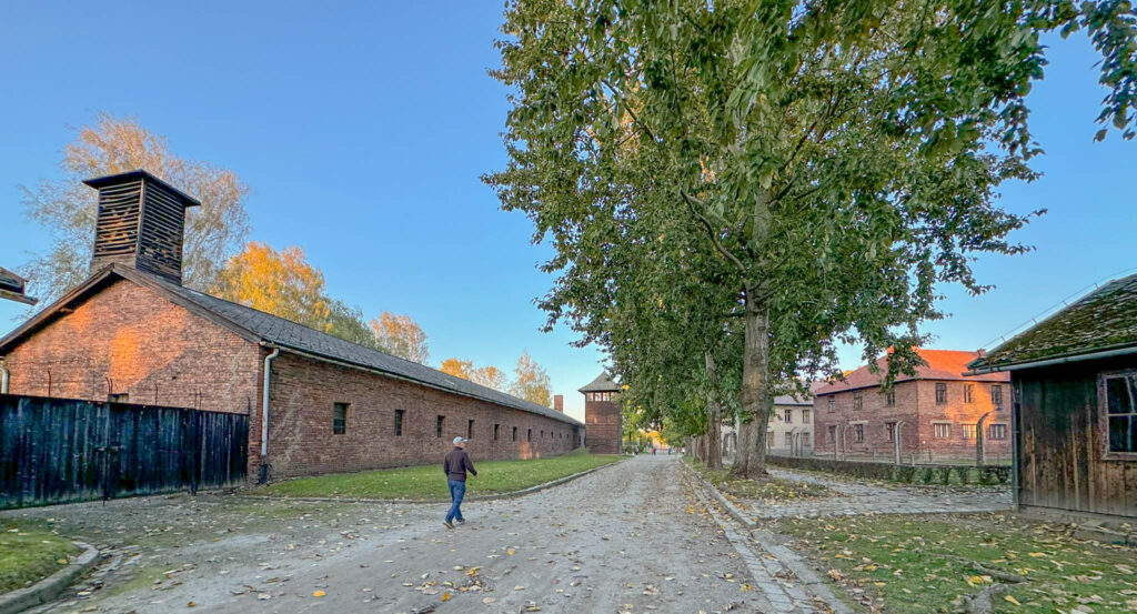 This image captures a scene from Auschwitz I, showing a long brick building with a wooden ventilation tower, which was likely used as a storage or administrative facility. In the background, a guard tower and barbed-wire fences are visible, emphasizing the camp’s strict security and confinement measures.