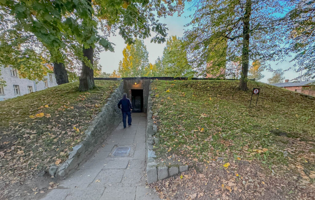 This image shows the entrance to the gas chamber and crematorium at Auschwitz I, a somber site of mass executions during the Holocaust. The pathway leads underground to a dimly lit doorway, blending into the grassy mound that conceals the structure. This unassuming yet chilling entrance marks the final moments for thousands of victims and now stands as a place of remembrance and historical reflection.