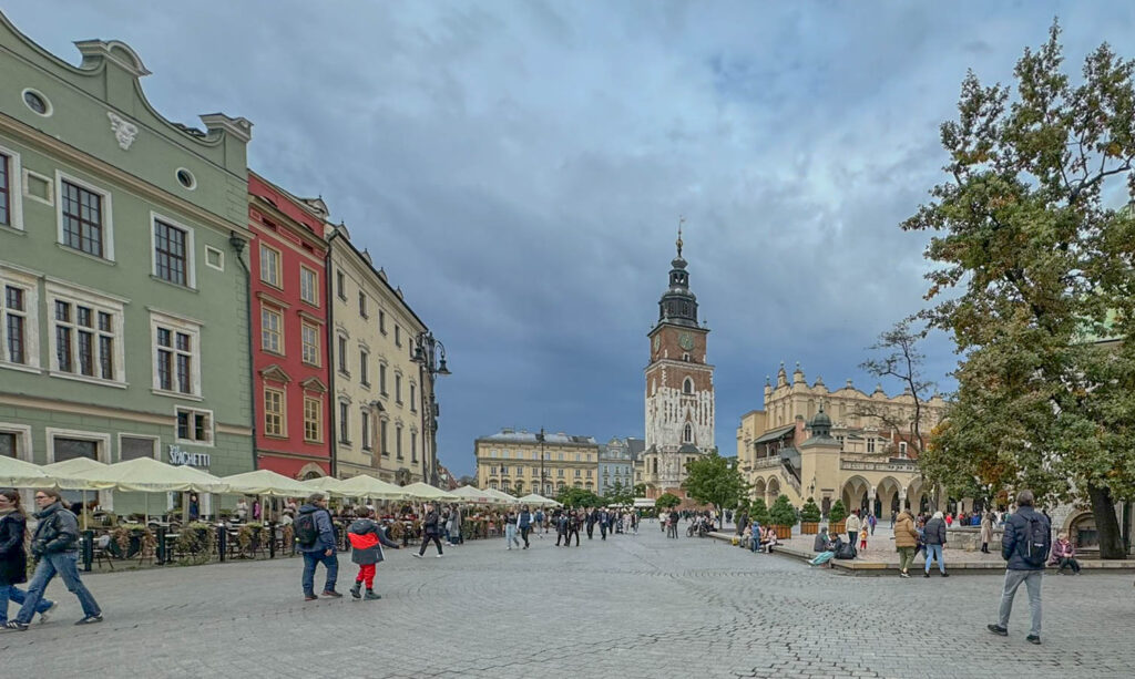 This image captures Kraków’s Main Market Square (Rynek Główny), the heart of the city’s historic Old Town. The tall tower in the center is the Town Hall Tower (Wieża Ratuszowa), the only remaining part of Kraków’s old town hall, which was demolished in the 19th century. To the right, the ornate building with arched arcades is the Cloth Hall (Sukiennice), a historic marketplace that has been a hub for trade since medieval times. The colorful townhouses and cafés on the left add to the square’s vibrant atmosphere, making it one of the most picturesque and lively spots in Kraków.