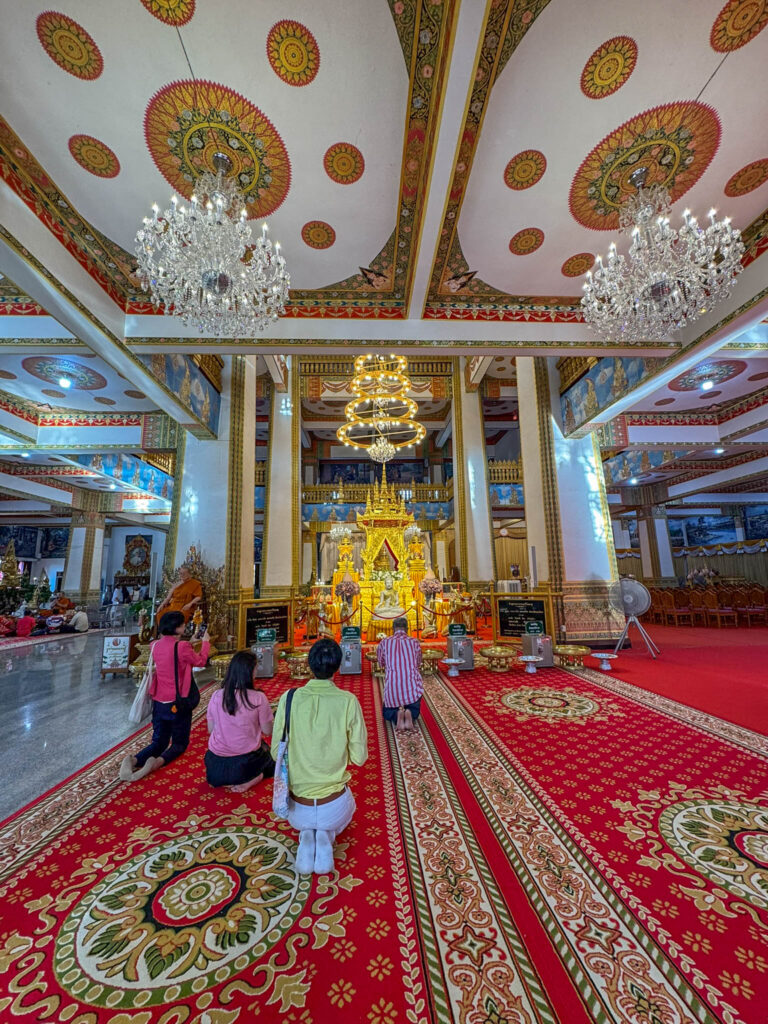 Visitors kneel in prayer beneath the chandeliers at Wat Nong Wang, with the main altar glowing at the center of it all.
